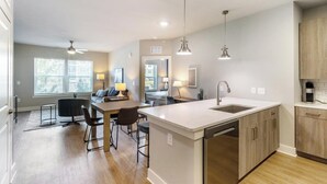 A modern kitchen with a central island, a dining area with a table and chairs, and a seating area with a sofa and a lamp. - Landing Furnished Apartments Near Point Washington State Forest (Santa Rosa Beach)
