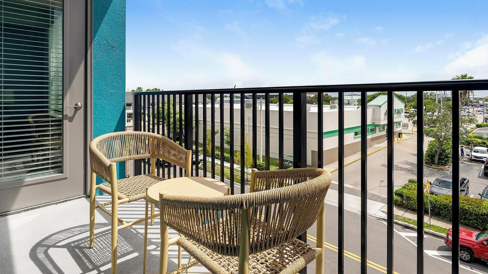 Un balcon avec des fauteuils en osier et une table, donnant sur un parking et un immeuble doté d’un toit végétalisé.