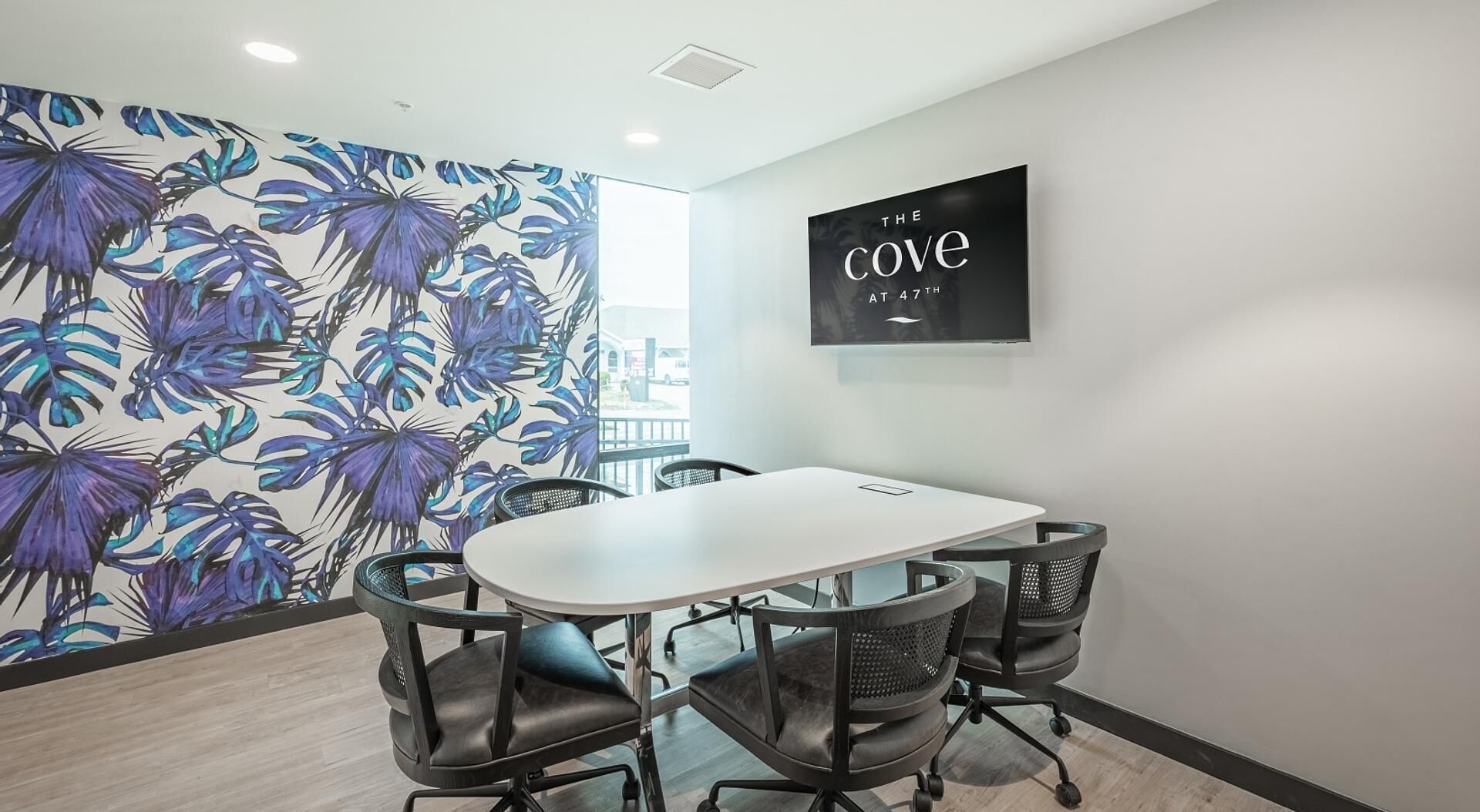 A conference room with a white table, four black office chairs, and a wall with a tropical leaf pattern. A black sign on the wall reads "THE COVE AT 47TH".