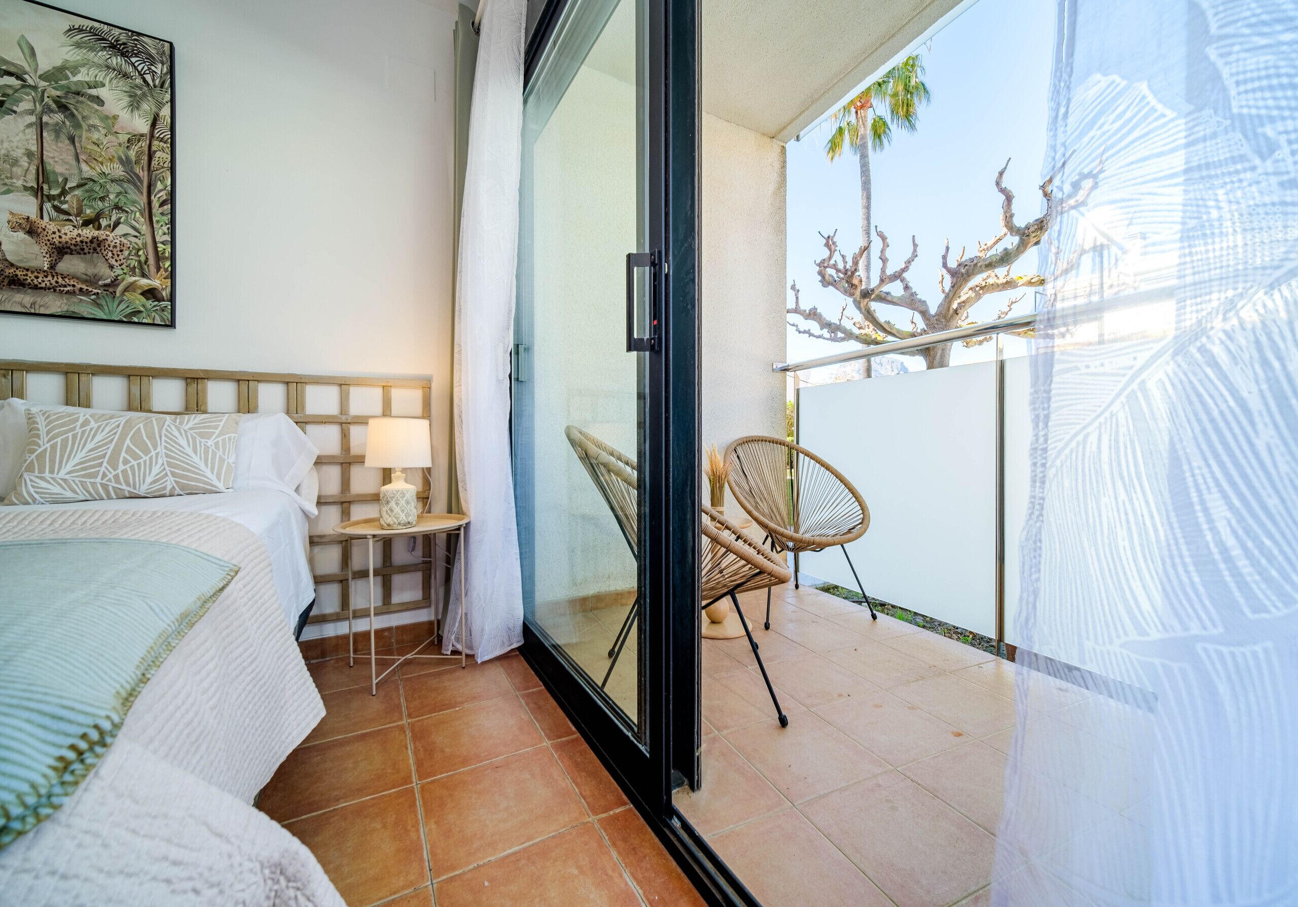 Quarto com cama, mesa de cabeceira, abajur e vista para uma varanda com cadeira e árvore.