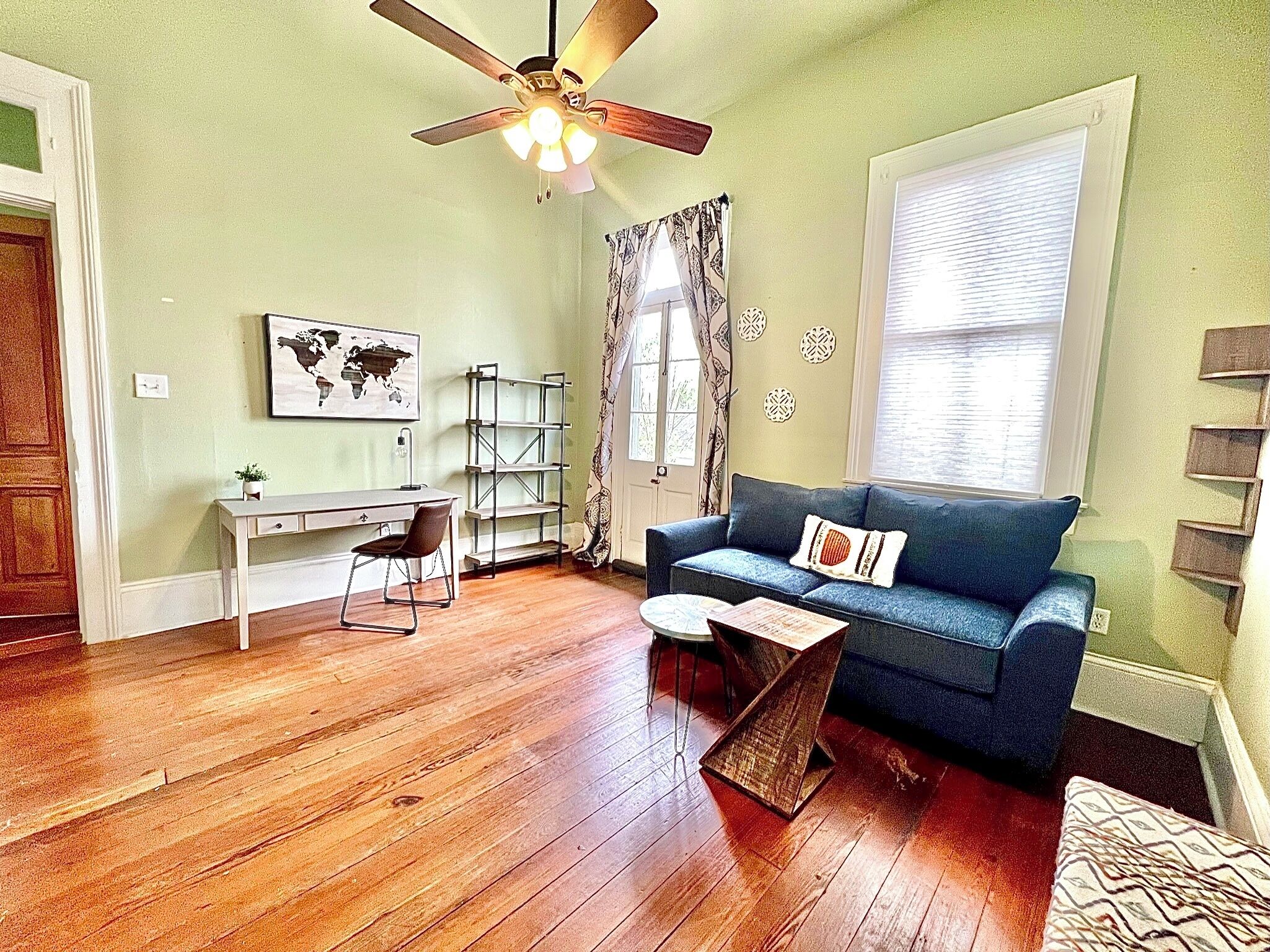 A living room with a wooden floor, a desk, a blue sofa, a ceiling fan, and a window with blinds.
