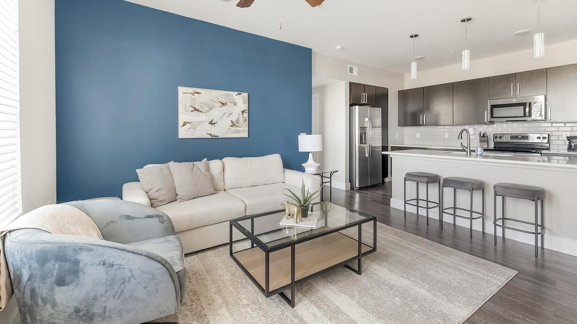A modern living room with a blue accent wall, a sofa, a coffee table, and a kitchen area with a refrigerator, microwave, and bar stools.