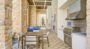 Une terrasse extérieure avec des murs en pierre, une table en bois, des chaises, un canapé bleu et un barbecue encastré.