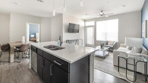 A modern kitchen with a central island, a dining area with a table and chairs, a living room with a sofa and a coffee table, and a ceiling fan. - Landing Furnished Apartments Allen - Twin Creeks (Allen)