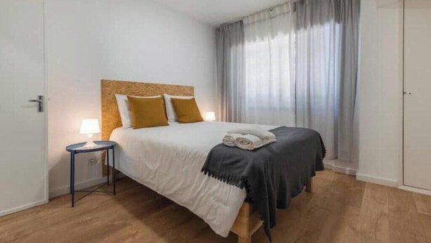 A hotel room with a bed, a side table with a lamp, a window with curtains, and a door. - Village Carcavelos II - Refuge with private backyard 7 minutes from the beach (Carcavelos)