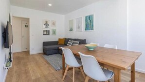 A modern living room with a wooden dining table, grey sofa, and a TV mounted on the wall. - Village Carcavelos II - Refuge with private backyard 7 minutes from the beach (Carcavelos)