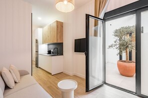 Egy modern nappali, melyben található egy kanapé, egy kis, kerek asztal, egy konyhasarok fa szekrényekkel, egy síkképernyős televízió, valamint egy tolóüveg-ajtó, amely a kültéri teraszra nyílik, ahol egy virágtartóban lévő növény áll.