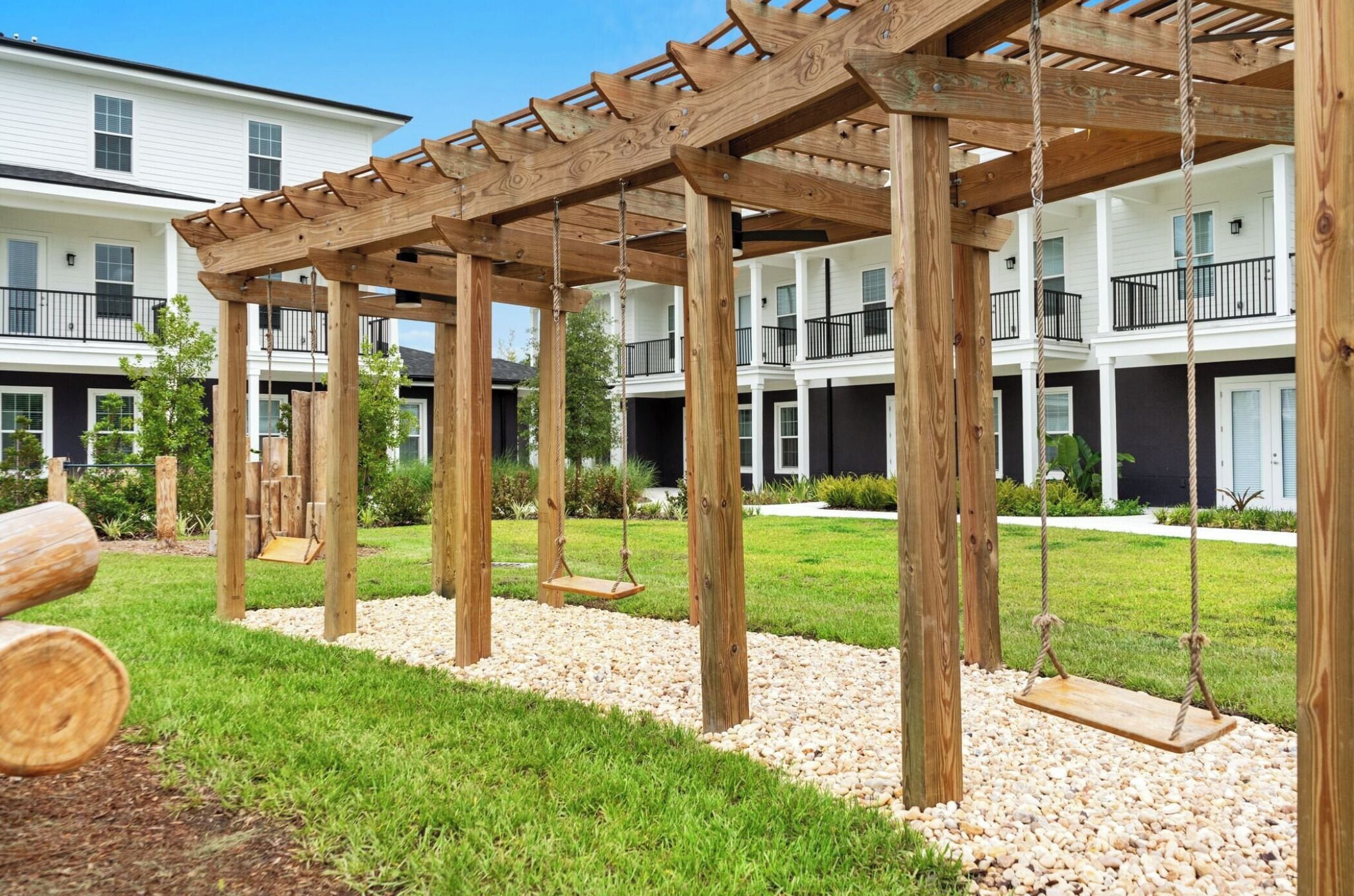 Eine hölzerne Pergola mit Schaukeln, umgeben von einem Kiesweg und viel Grün, mit mehrstöckigen Gebäuden im Hintergrund.