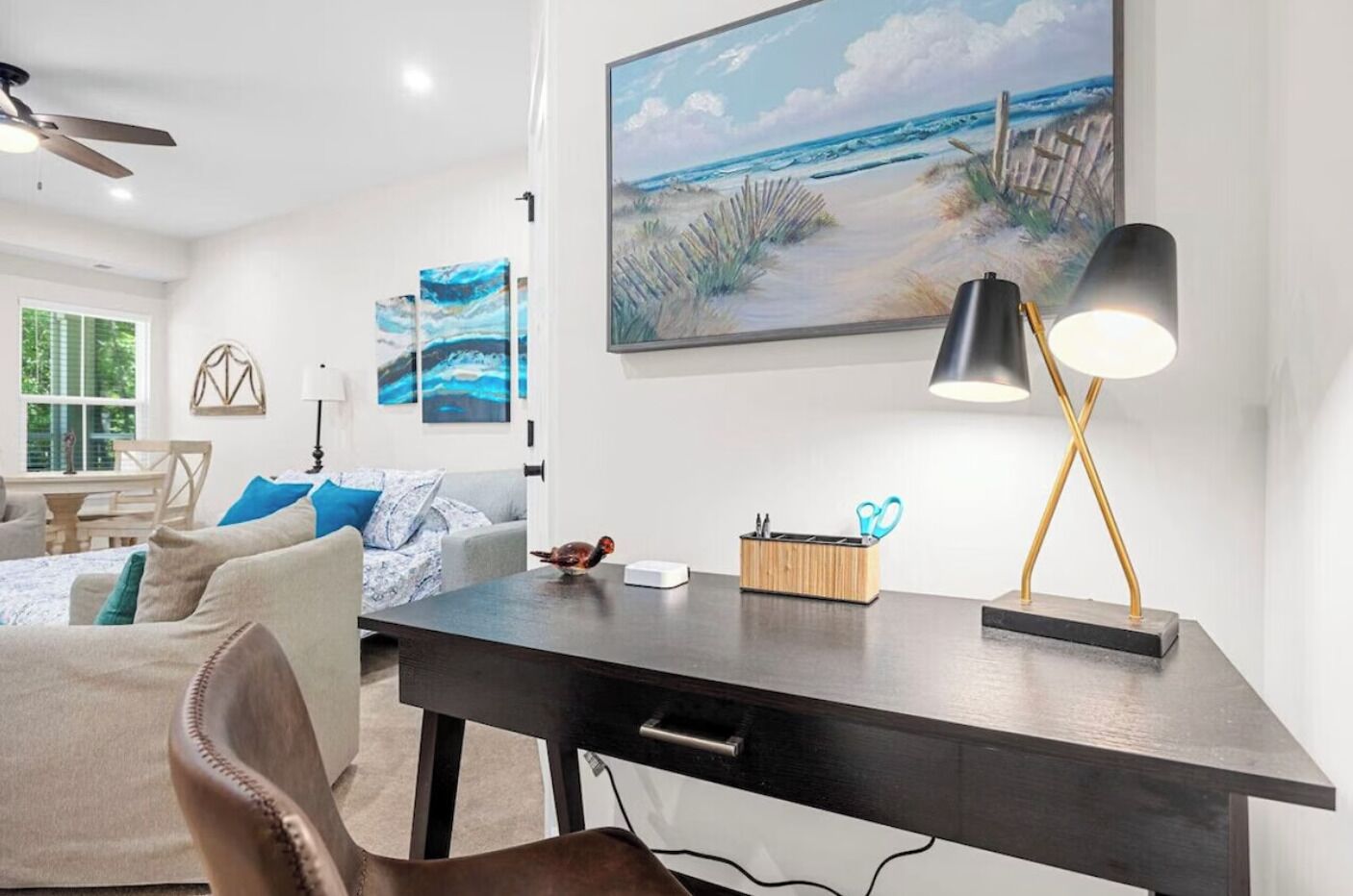 A modern living room with a dark wooden desk, a painting of a beach, and a ceiling fan.