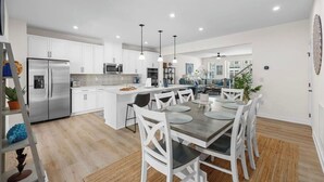 A modern kitchen with white cabinets, stainless steel appliances, and a dining area with white chairs and a wooden table. - Luxe Dye Estates Townhome @ Barefoot - Golf, Pool, Hot Tub (North Myrtle Beach)