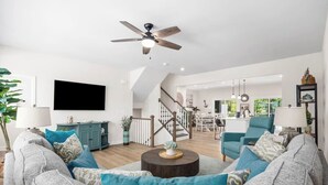 A spacious living area with a large sofa, a coffee table, a TV, and a staircase leading to a dining area. - Luxe Dye Estates Townhome @ Barefoot - Golf, Pool, Hot Tub (North Myrtle Beach)