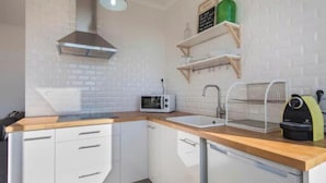 Een kleine keuken met witte kastjes, een magnetron, een spoelbak, een waterkoker en een hangende rek.