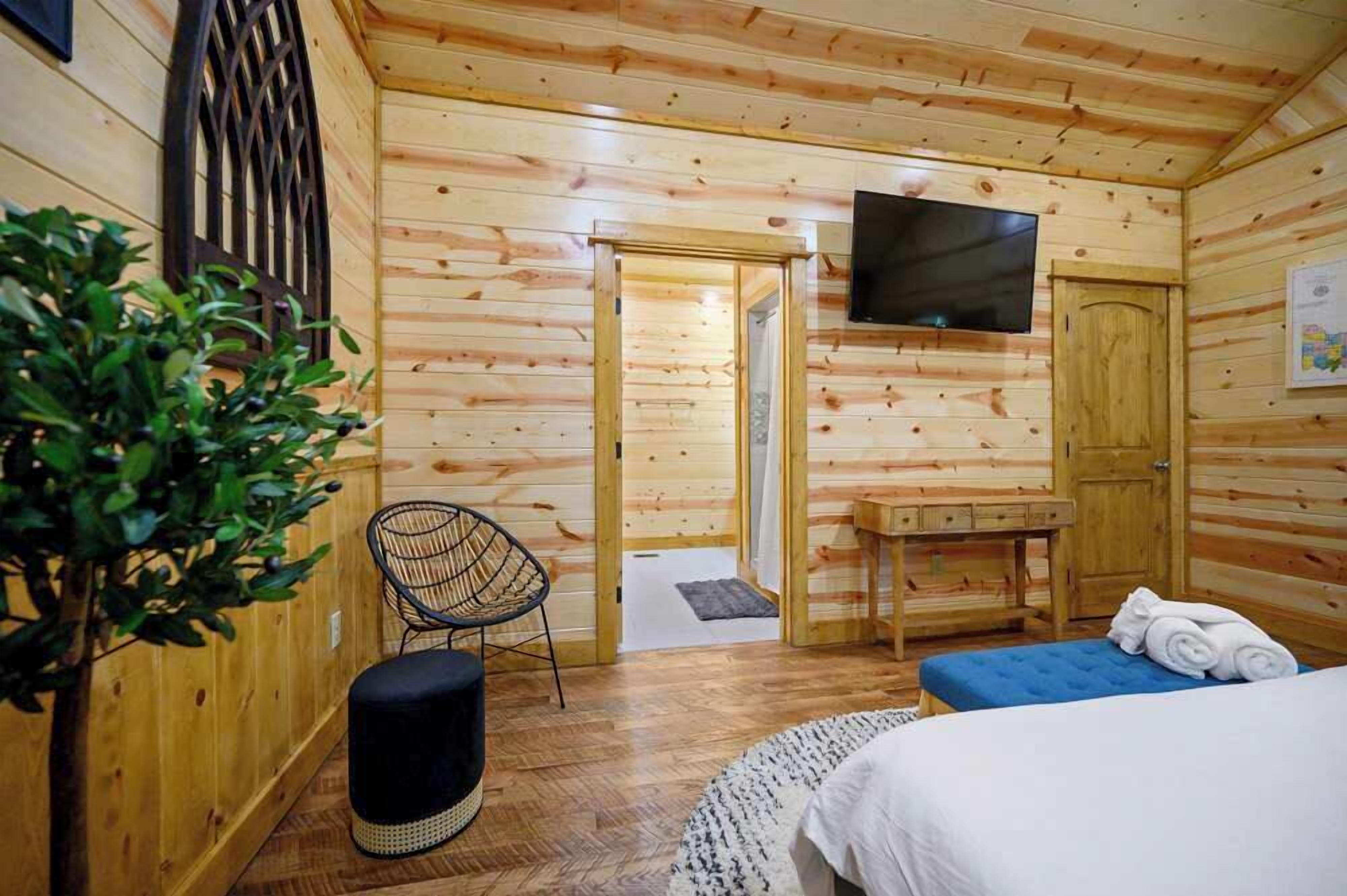 Ein Zimmer in einer Holzhütte mit einem Bett, einem Fernseher, einem Sessel, einem kleinen Tisch und einer Tür zu einem anderen Raum.