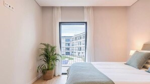 Makuuhuoneessa on sänky, ruukussa oleva kasvi, ikkuna, josta on näkymä rakennuksiin, ja seinässä valokytkin.
