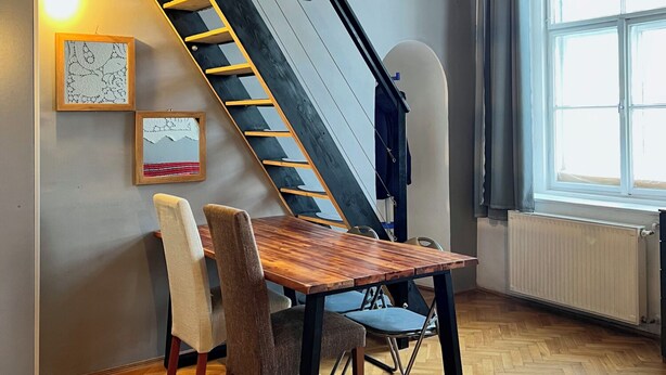 A dining area with a wooden table and chairs, a staircase, and a large window. - Cozy Loft Retreat by Chain Bridge – Stunning Danube & Parliament Views (Budapest)