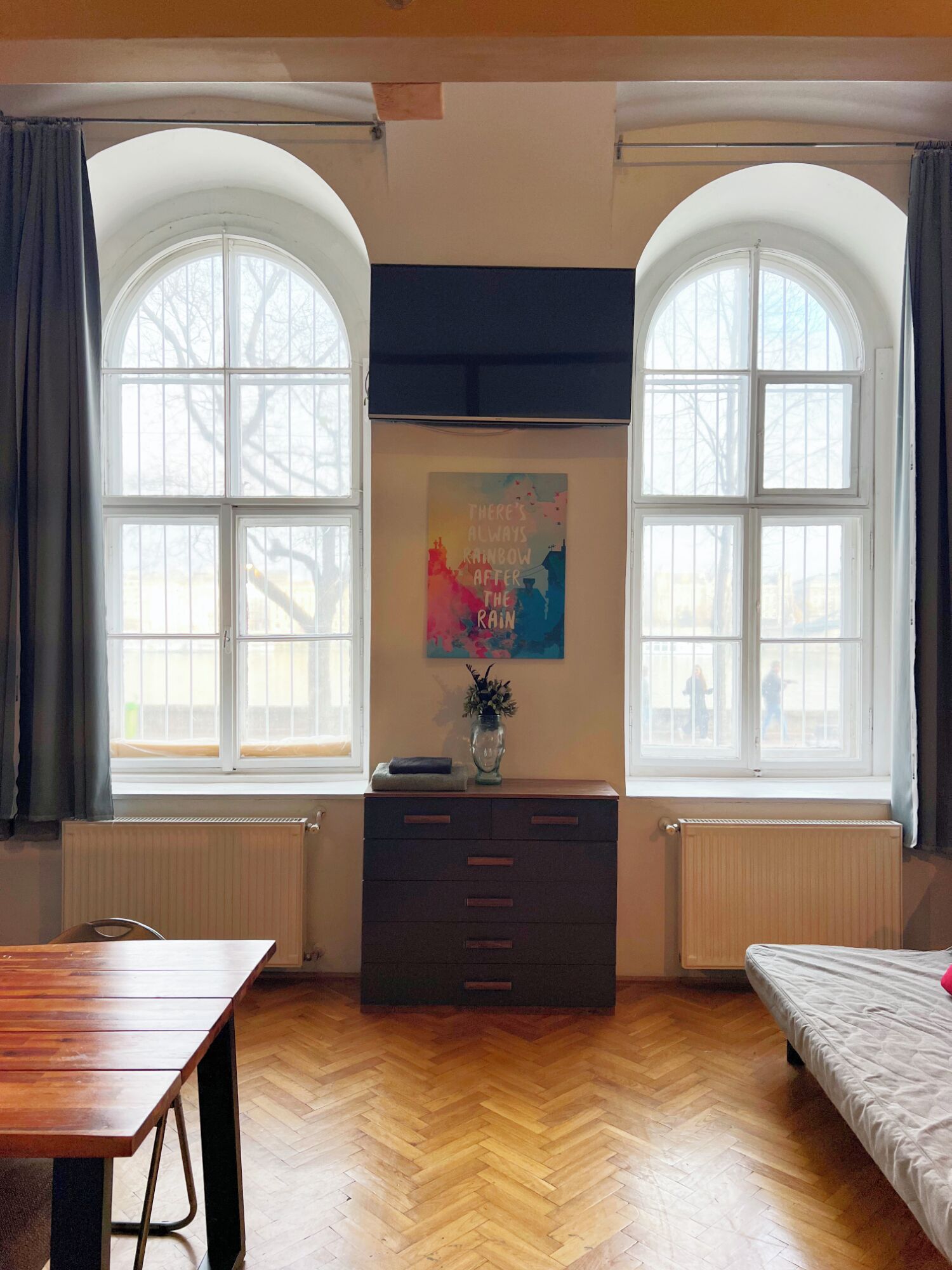 A room with two large arched windows, a wooden herringbone floor, a dresser with drawers, a table, and a bed.