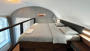 A modern bedroom with a curved ceiling, a bed with white linens, a cityscape painting, a standing fan, and a window with curtains. - Cozy Loft Retreat by Chain Bridge – Stunning Danube & Parliament Views (Budapest)