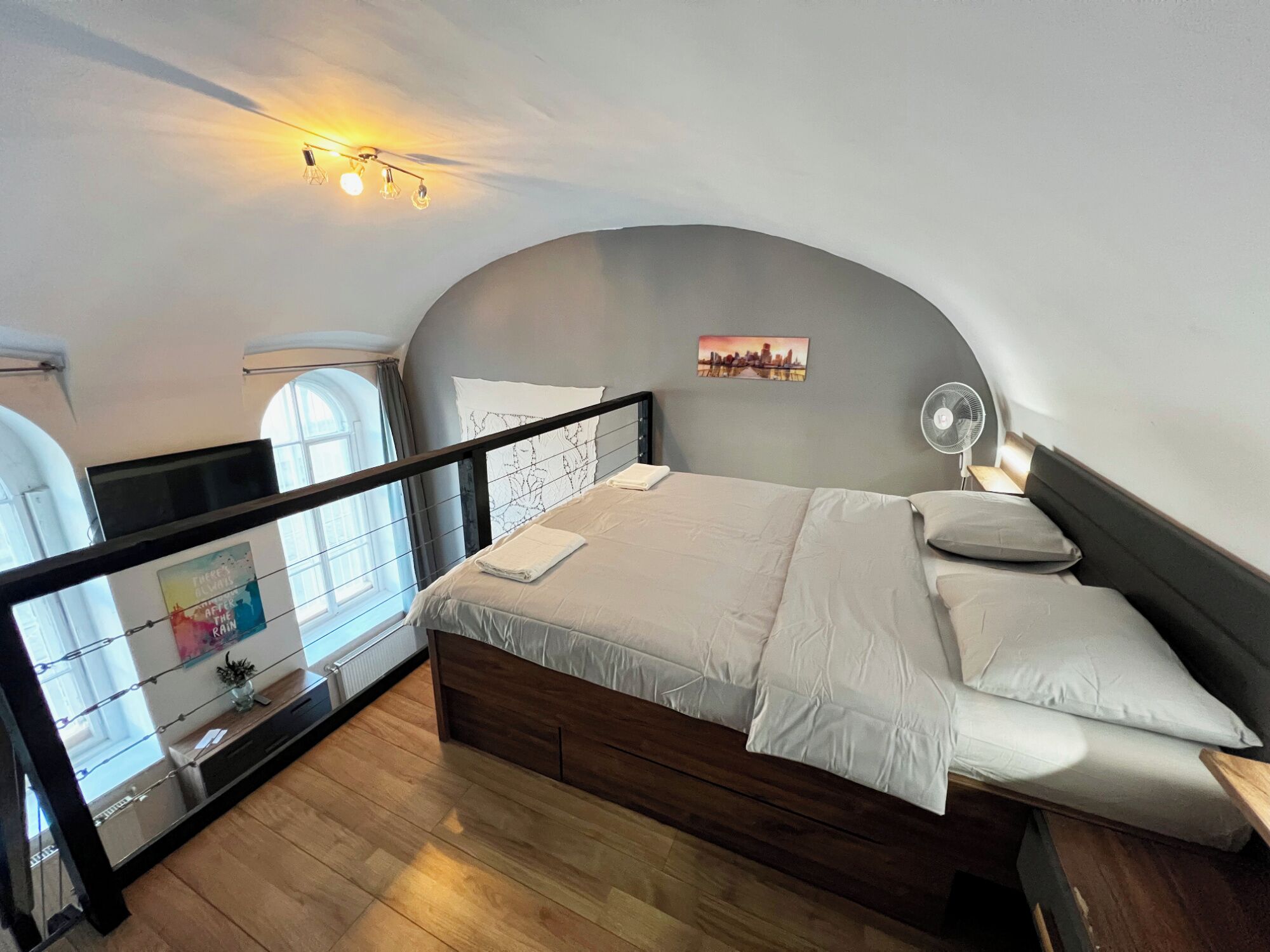 A bedroom with a wooden bed, a white headboard, a cityscape picture, a bedside table with a lamp, and a window with curtains.