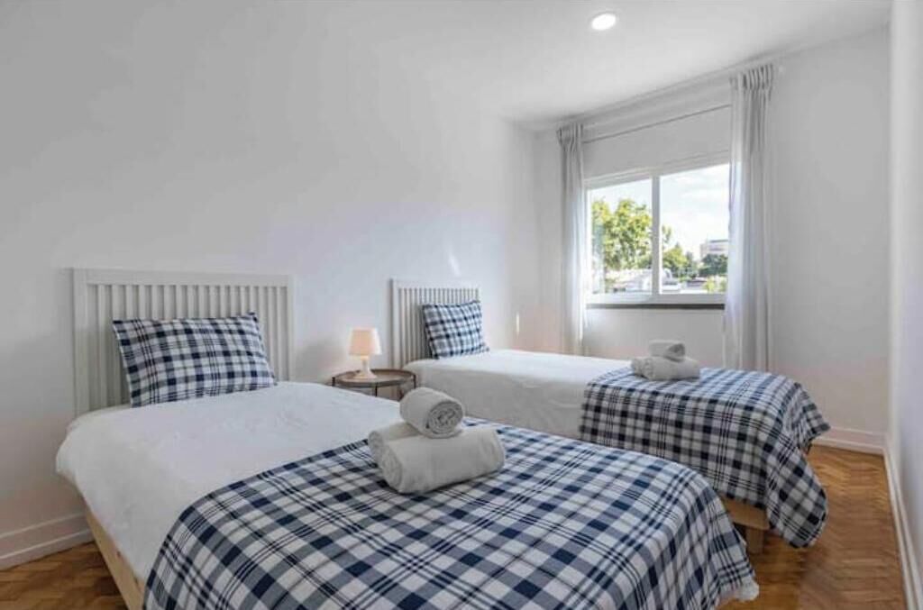 A hotel room with two beds, white bedding, a window with curtains, and a radiator.