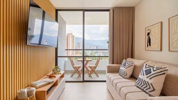 Ein modernes Wohnzimmer mit einer Couch, einem an der Wand befestigten Fernseher, einem Balkon mit Tisch und Stühlen sowie Blick auf die Stadt.