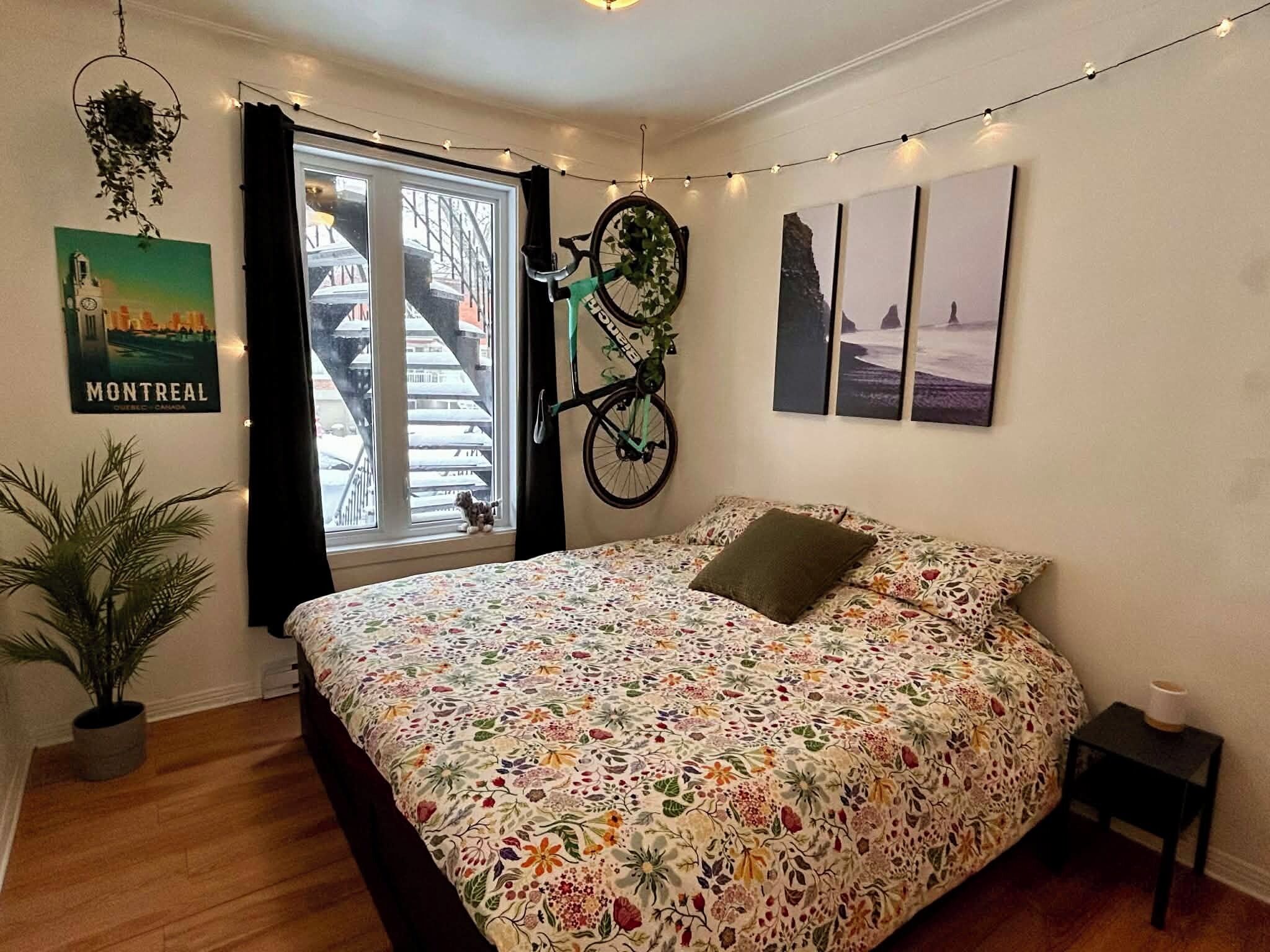 A bedroom with a floral bedspread, a bicycle hanging on the wall, a poster of Montreal, and a string of lights.