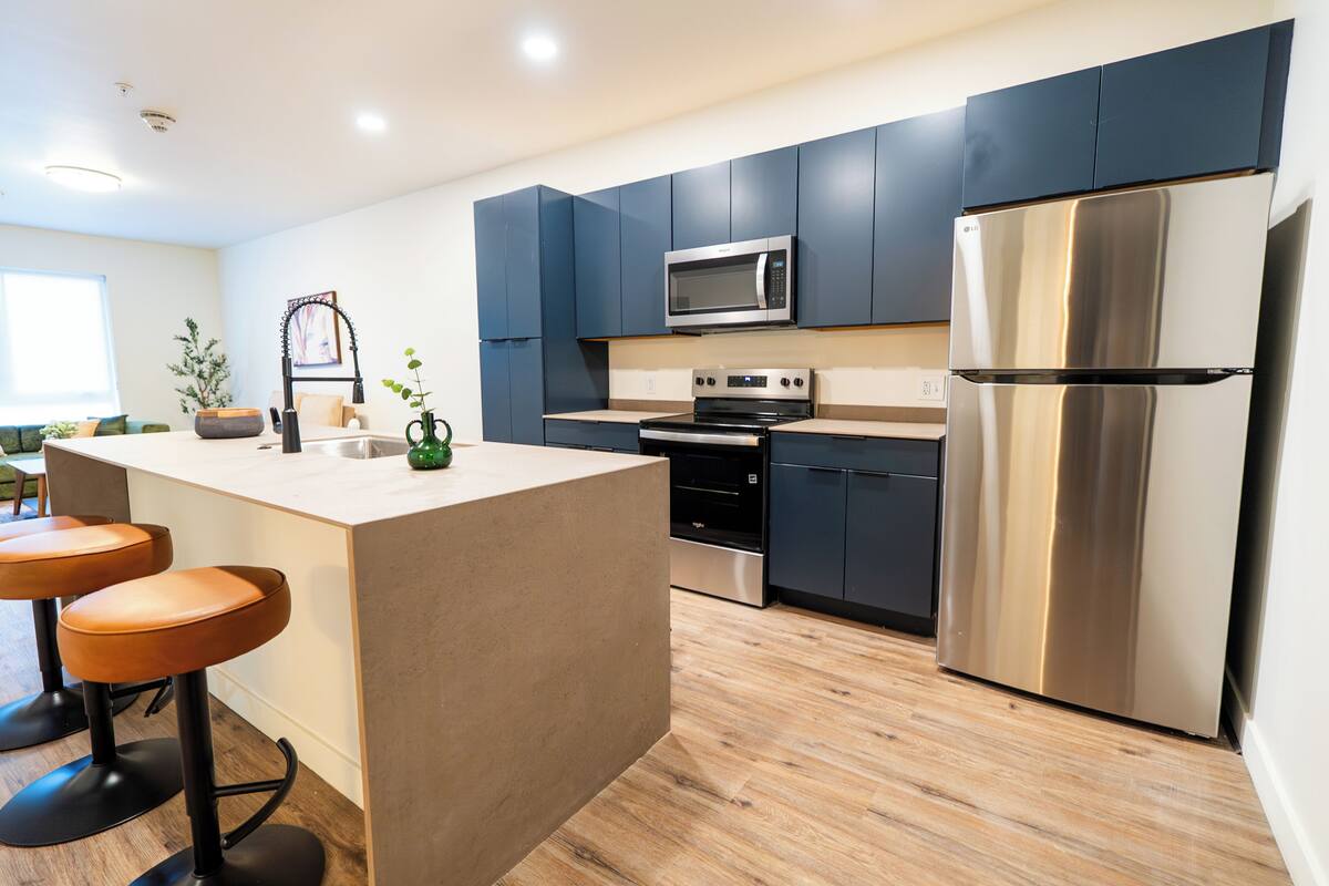 A modern kitchen with dark cabinets, stainless steel appliances, a central island with stools, and a wooden floor.