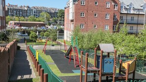 Ein Spielplatz mit Schaukeln und Klettergerüsten, umgeben von einem grünen Zaun und einem Backsteingebäude im Hintergrund.