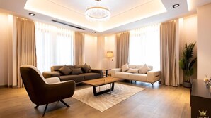 Una sala de estar moderna con un sofá, un sillón y una mesa de centro, una araña y ventanas grandes con cortinas.