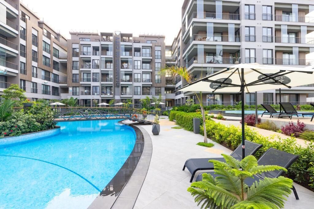 Una piscina rodeada de sillas de descanso y follaje, con edificios de apartamentos modernos al fondo.