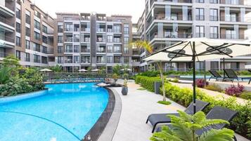 Una piscina rodeada de sillas de descanso y follaje, con edificios de apartamentos modernos al fondo.