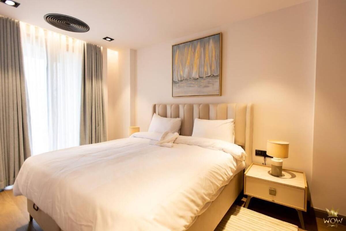 Egy szállodai szoba, amelyben egy nagy ágy, egy éjjeliszemélyke lámpával, egy vitorláshajókat ábrázoló festmény és egy mennyezeti ventilátor található.