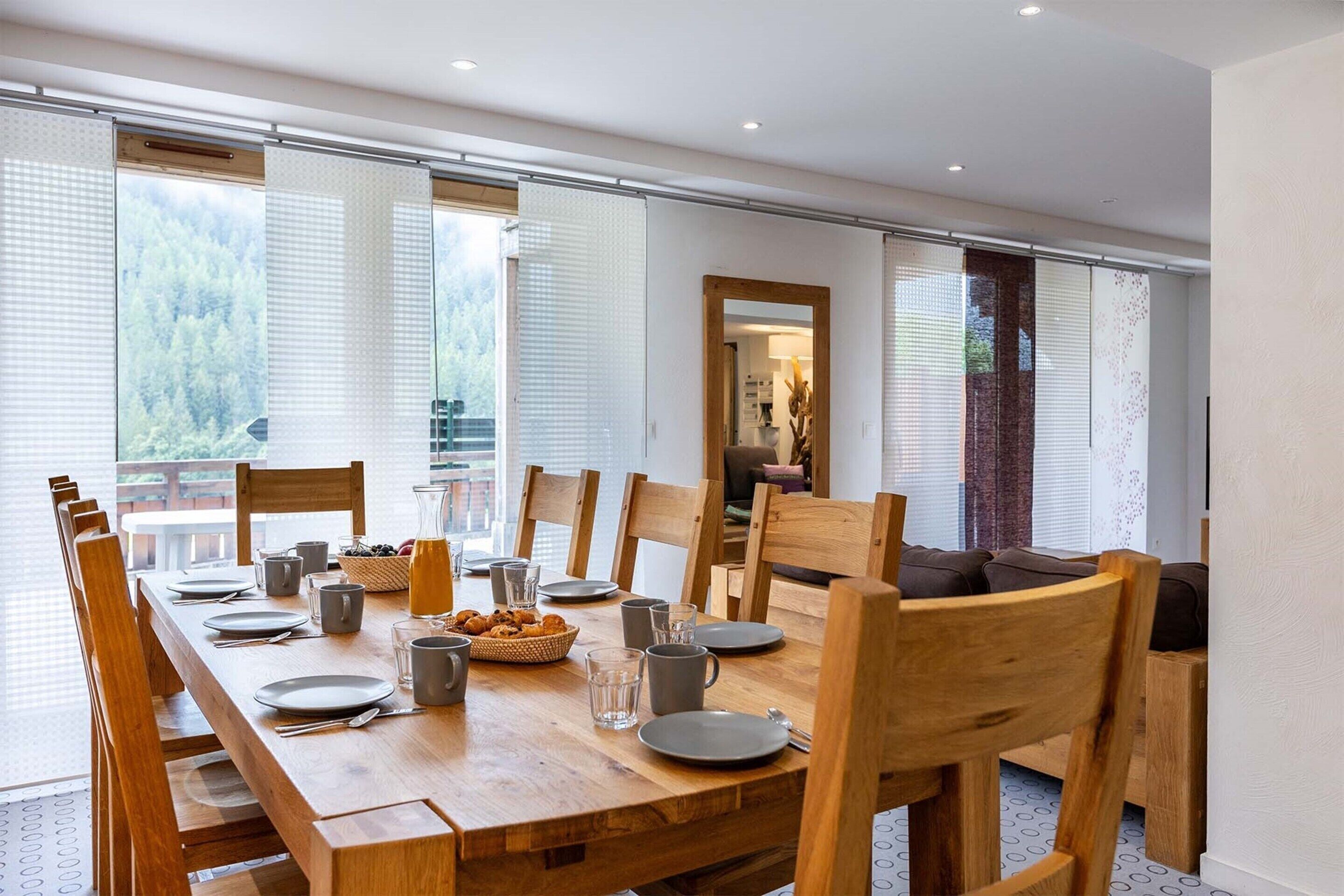 A dining area with a long wooden table, chairs, and a view of a forest through large windows.