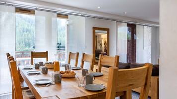 A dining area with a long wooden table, chairs, and a view of a forest through large windows.
