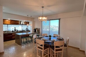 A modern kitchen with a large island, dining area with chairs, and a large window with a city view. - Lugar hermoso, cómodo y céntrico. ¿Algo más? (Cabo San Lucas)
