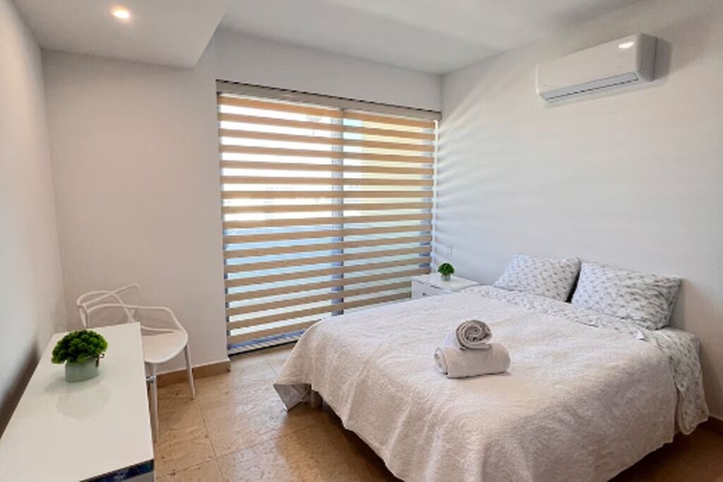 A bedroom with a bed, a bedside table, a chair, a window with blinds, and an air conditioning unit.