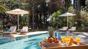 Ambiente à beira da piscina com cadeiras de descanso, guarda-sóis e uma bandeja de frutas e sucos.
