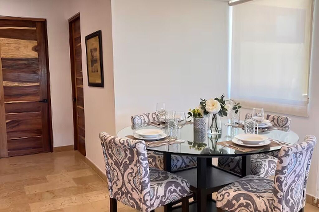 Een eetgedeelte met een ronde glazen tafel, gestreept beklede stoelen en een vaas met bloemen.