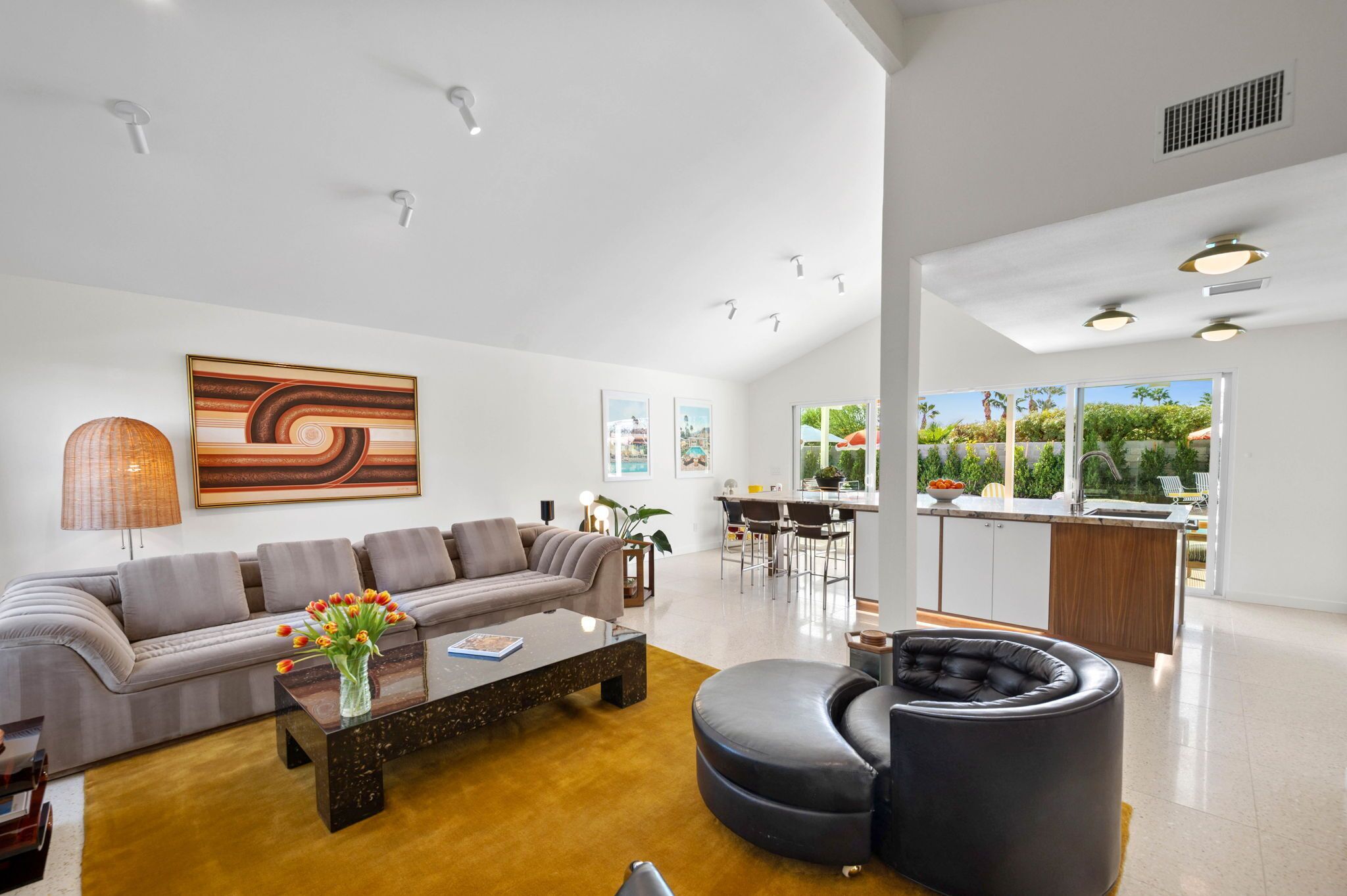 A modern living room with a sofa, coffee table, and ottoman, a dining area with chairs, and a kitchen with a bar.