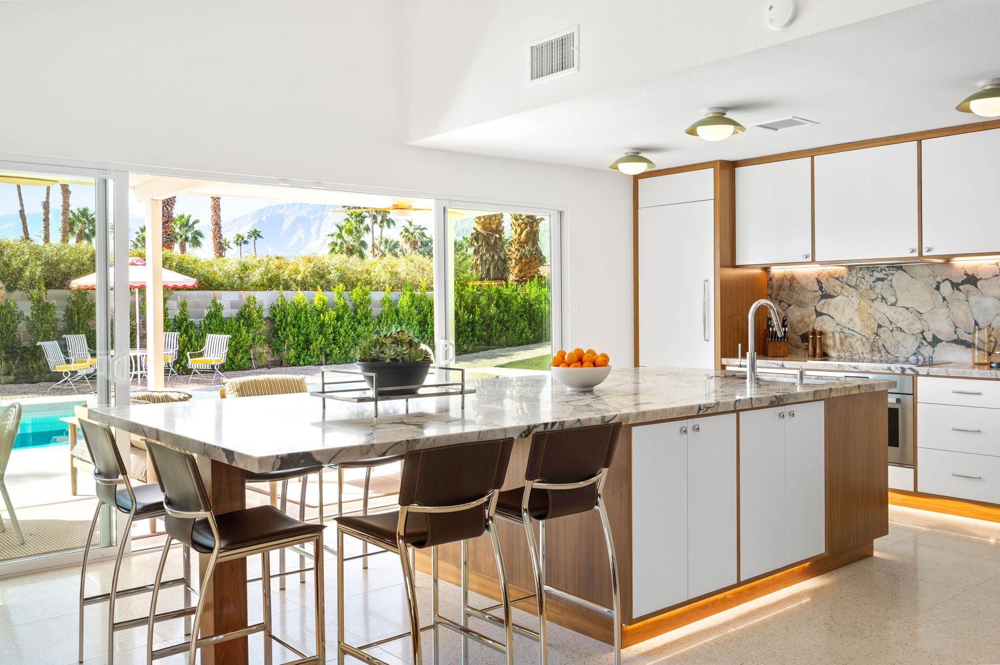 A modern kitchen with a large island, bar stools, a marble countertop, and a view of a pool and greenery.