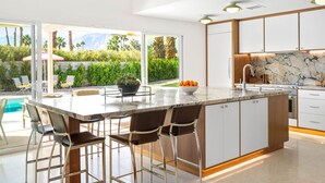 Une cuisine moderne dotée d’un grand îlot central, de tabourets de bar, d’un plan de travail en marbre et offrant une vue sur une piscine et un espace verdoyant.