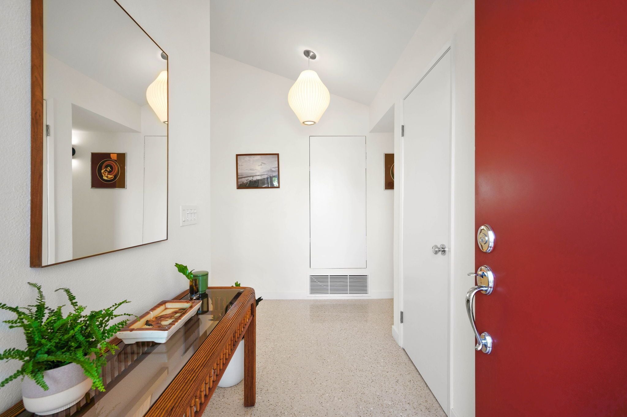 Un couloir avec un banc en bois, une plante en pot, un miroir et une porte rouge.