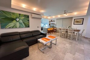 Ein Wohnzimmer mit schwarzem Sofa, Couchtisch, Essbereich mit Stühlen, einer dahinterliegenden Küche und einem Deckenventilator.