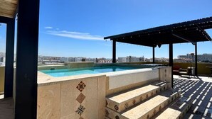 Uma área de piscina no terraço com escadas descendo até a piscina, uma pérgola e vista clara do horizonte da cidade.