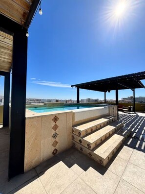 Ein Poolbereich auf dem Dach mit Stufen zum Pool, einer Pergola und freiem Blick auf die Stadtlandschaft.