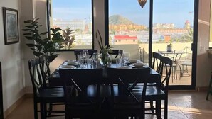 Uma sala de jantar com mesa posta para quatro pessoas, janelas amplas com vista para a cidade e um vaso de planta.
