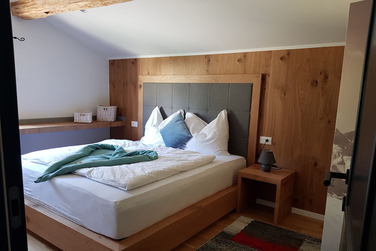 A bedroom with a wooden headboard, a bed with white bedding, a bedside table with a lamp, and a small shelf with a basket.