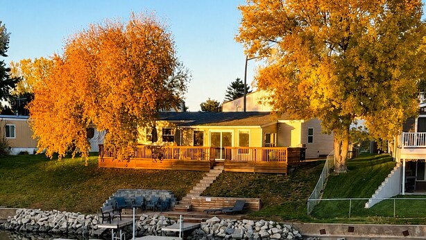 Una propiedad junto al lago con una superficie acuática reflectante, una terraza de madera y un sendero de piedra que conduce a un edificio con porche.