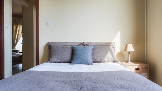 Ein ordentlich bezogenes Bett mit einer grauen Decke, ein Nachttisch aus Holz mit einer Lampe und ein Spiegel an der Wand.