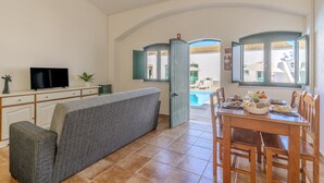 En stue med sofa, fjernsyn, spisebord med stole og udsigt til et udendørs swimmingpool.