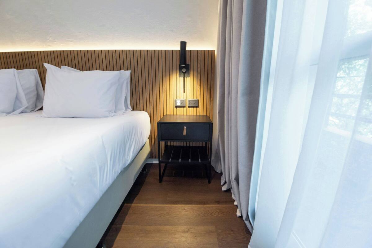 Habitación de hotel con cama, mesita de noche con lámpara, paneles de madera en la pared y ventana con cortinas.